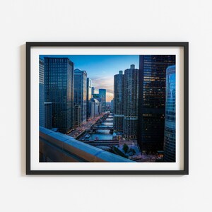 Pode incluir: Uma vista panorâmica de um horizonte urbano ao entardecer. A imagem mostra uma série de edifícios altos com janelas de vidro, que refletem o céu azul e o pôr do sol. Um rio atravessa a cidade, com pontes que conectam os edifícios de ambos os lados.