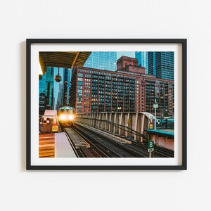 May include: A train pulls into a station in a city with tall buildings. The train is lit up with yellow lights and the station is made of concrete and metal.