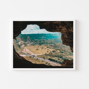 May include: A view of a lush green valley from inside a cave opening. The valley is filled with trees and a river winding through it. The cave opening is dark and rocky.