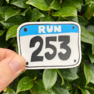 May include: A white sticker with a blue top section that reads "RUN" above the number "253" in bold black font. The sticker has a black border and is held against a background of green leaves.
