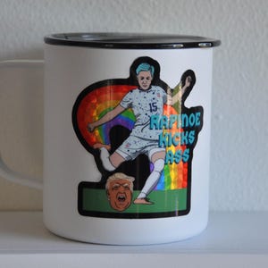 K&ouml;nnte beinhalten: Eine wei&szlig;e Emailletasse mit schwarzem Rand und Henkel. Ein Aufkleber auf der Tasse zeigt eine Karikatur eines Fu&szlig;ballspielers, der einen Fu&szlig;ball tritt, mit einem Regenbogenhintergrund und dem Text "Rapinoe Kicks Ass".