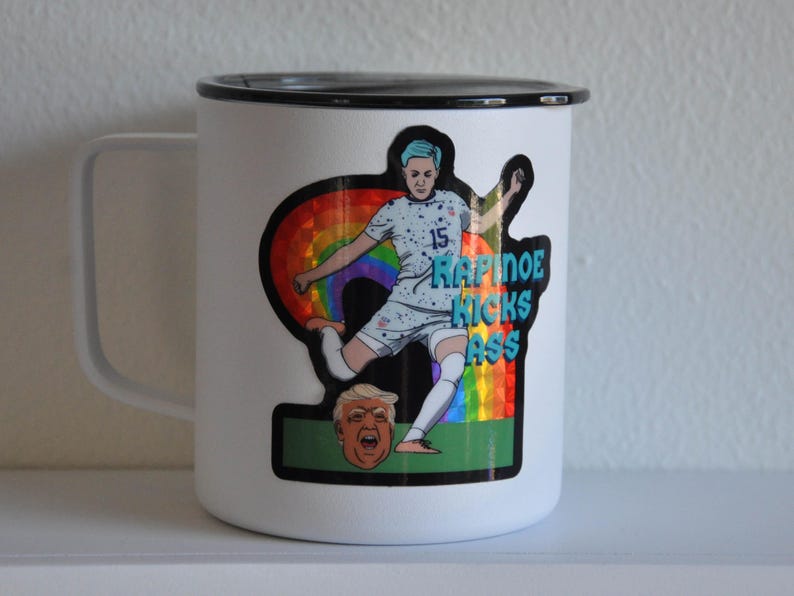 K&ouml;nnte beinhalten: Eine wei&szlig;e Emailletasse mit schwarzem Rand und Henkel. Ein Aufkleber auf der Tasse zeigt eine Karikatur eines Fu&szlig;ballspielers, der einen Fu&szlig;ball tritt, mit einem Regenbogenhintergrund und dem Text "Rapinoe Kicks Ass".