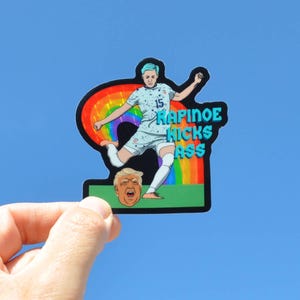 K&ouml;nnte beinhalten: Ein Aufkleber mit einer Cartoon-Illustration einer Fu&szlig;ballspielerin, die einen Fu&szlig;ball tritt. Die Spielerin tr&auml;gt ein blau-wei&szlig;es Trikot mit der Nummer 15. Die Spielerin tritt einen Fu&szlig;ball in Richtung eines Regenbogens. Der Fu&szlig;ball ist eine Cartoon-Darstellung eines Menschenkopfes. Der Text "RAPINOE KICKS ASS" ist in blauen Buchstaben auf dem Aufkleber geschrieben.