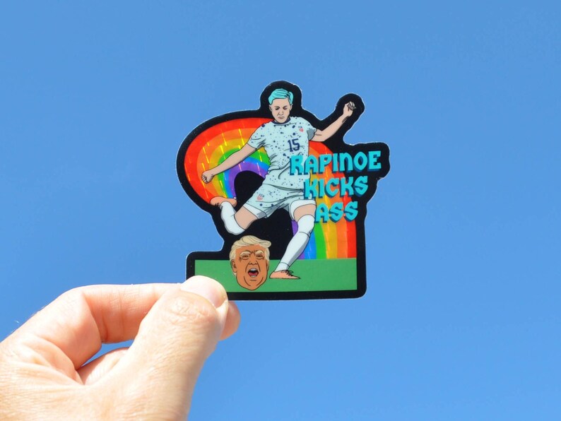 K&ouml;nnte beinhalten: Ein Aufkleber mit einer Cartoon-Illustration einer Fu&szlig;ballspielerin, die einen Fu&szlig;ball tritt. Die Spielerin tr&auml;gt ein blau-wei&szlig;es Trikot mit der Nummer 15. Die Spielerin tritt einen Fu&szlig;ball in Richtung eines Regenbogens. Der Fu&szlig;ball ist eine Cartoon-Darstellung eines Menschenkopfes. Der Text "RAPINOE KICKS ASS" ist in blauen Buchstaben auf dem Aufkleber geschrieben.