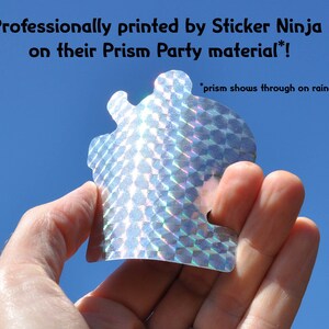 K&ouml;nnte beinhalten: Ein silberner holografischer Aufkleber mit einem geometrischen Muster. Der Aufkleber wird in einer Hand vor einem blauen Himmel gehalten. Der Text "Professionally printed by Sticker Ninja on their Prism Party material!" befindet sich &uuml;ber dem Aufkleber. Der Text "*prism shows through on rainbow" befindet sich unter dem Aufkleber.
