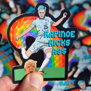 K&ouml;nnte beinhalten: Ein Aufkleber mit einer Cartoon-Illustration einer weiblichen Fu&szlig;ballspielerin, die einen Fu&szlig;ball in Richtung des Kopfes eines Mannes tritt. Die Fu&szlig;ballspielerin tr&auml;gt ein blaues und wei&szlig;es Fu&szlig;balltrikot mit der Nummer 15 auf der Vorderseite. Der Aufkleber hat einen Regenbogenhintergrund und den Text "RAPINOE KICKS ASS" in blauer und wei&szlig;er Schrift.