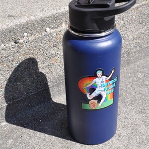 K&ouml;nnte beinhalten: Eine blaue Edelstahl-Wasserflasche mit schwarzem Deckel und einem Aufkleber einer Frau, die einen Fu&szlig;ball tritt, mit dem Text "Rapinoe Kicks Ass" darauf.
