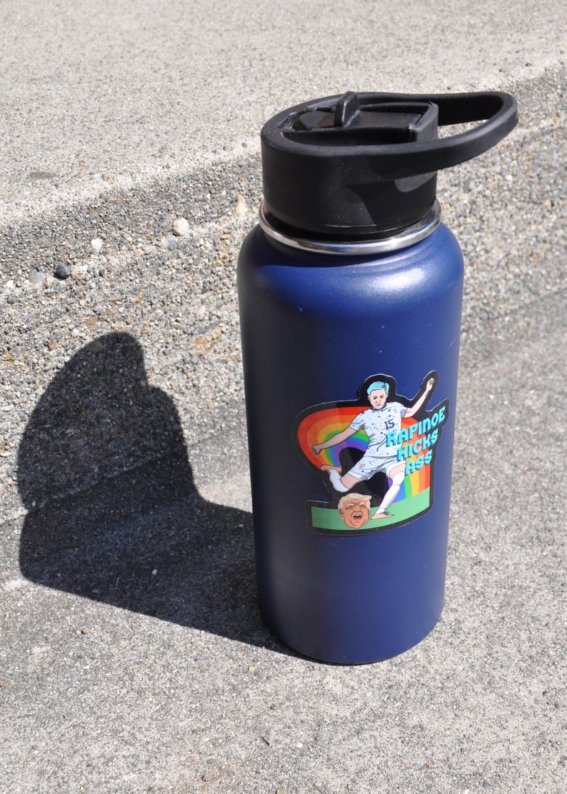 K&ouml;nnte beinhalten: Eine blaue Edelstahl-Wasserflasche mit schwarzem Deckel und einem Aufkleber einer Frau, die einen Fu&szlig;ball tritt, mit dem Text "Rapinoe Kicks Ass" darauf.