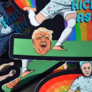 K&ouml;nnte beinhalten: Ein Aufkleber mit einem Cartoon-Bild eines Fussballspielers, der einen Fussball tritt. Der Spieler tr&auml;gt ein weiss-blaues Trikot mit den Buchstaben "USA" auf der Brust. Der Fussball ist ein Cartoon-Bild von Donald Trumps Kopf. Der Aufkleber hat einen Regenbogen-Hintergrund und den Text "KICKS ASS" in blauen Buchstaben.