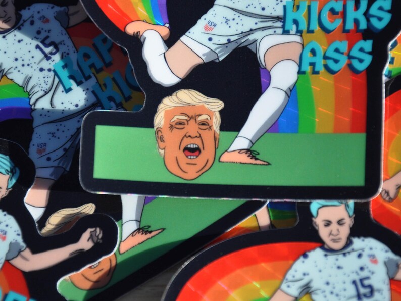 K&ouml;nnte beinhalten: Ein Aufkleber mit einem Cartoon-Bild eines Fussballspielers, der einen Fussball tritt. Der Spieler tr&auml;gt ein weiss-blaues Trikot mit den Buchstaben "USA" auf der Brust. Der Fussball ist ein Cartoon-Bild von Donald Trumps Kopf. Der Aufkleber hat einen Regenbogen-Hintergrund und den Text "KICKS ASS" in blauen Buchstaben.