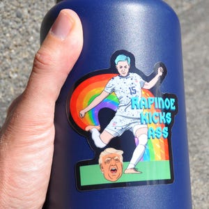 K&ouml;nnte beinhalten: Eine blaue Wasserflasche mit einem Aufkleber einer Fu&szlig;ballspielerin, die einen Fu&szlig;ball tritt. Der Aufkleber hat einen Regenbogen und den Text "RAPINOE KICKS ASS".