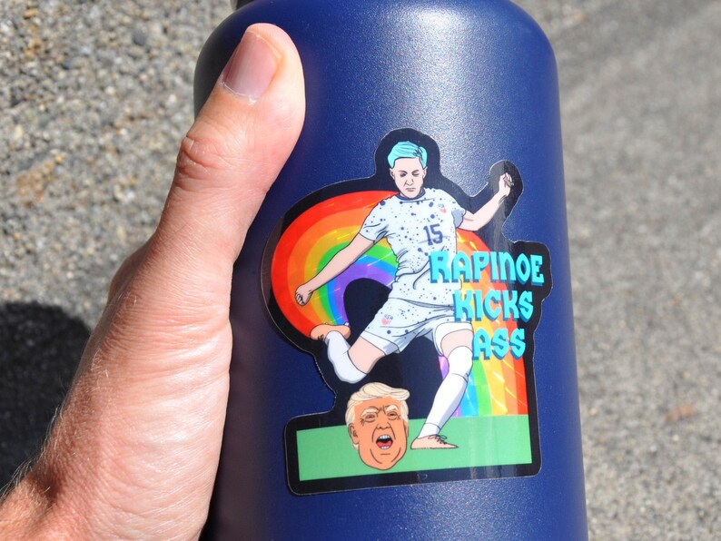 K&ouml;nnte beinhalten: Eine blaue Wasserflasche mit einem Aufkleber einer Fu&szlig;ballspielerin, die einen Fu&szlig;ball tritt. Der Aufkleber hat einen Regenbogen und den Text "RAPINOE KICKS ASS".