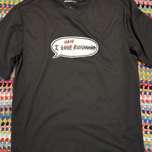 May include: Black short-sleeved t-shirt with a speech bubble graphic. The speech bubble has the words "HATE" crossed out in red, and "I LOVE RUNNING" in white text. The shirt is made of a lightweight material.
