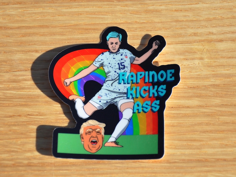 Colorful illustration of Megan Rapinoe kicking the head of a jerk with a sweeping rainbow behind her which features a holographic, prism effect in direct light.