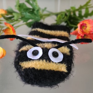 May include: A handmade plush bee with black and yellow stripes, button eyes, and pipe cleaner antennae. The bee is a whimsical design, possibly a toy or decorative item. The background includes colorful flowers.