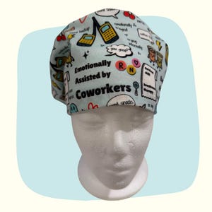 May include: A light blue scrub cap featuring a repeating pattern of cartoon pagers, cherries, lightning bolts, and speech bubbles. The text "Emotionally Assisted by Coworkers" is printed in black, alongside "RN" in a circle and various other small icons.