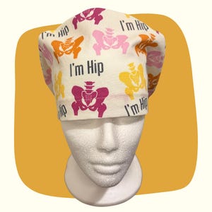 May include: A white surgical scrub cap with a repeating pattern of colorful hip bone illustrations in shades of pink, orange, and yellow. The words "I'm Hip" are printed in black. The cap is displayed on a white mannequin head.