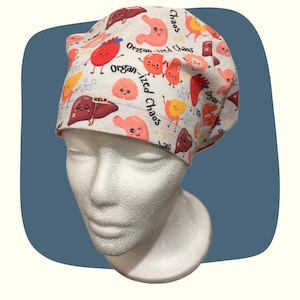 May include: A white surgical cap with a pattern of cartoon organs in red, yellow, and brown. The cap has the words "Organ-ized Chaos" and "Help" printed on it. The cap is designed to be worn in a medical setting.