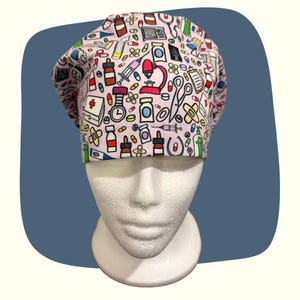 May include: A light pink medical scrub cap featuring a repeating pattern of medical tools and supplies. The pattern includes pills, syringes, stethoscopes, and thermometers. The cap is worn on a white mannequin head.