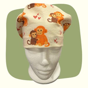 May include: A white and beige scrub cap featuring a repeating pattern of orange monkeys and pink hearts. The cap has a folded brim and is designed to fit the head. The monkeys are hugging each other.