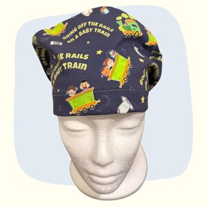 May include: Navy blue surgical scrub cap with a repeating pattern of a cartoon train, children, stars, and the text "GOING OFF THE RAILS ON A BABY TRAIN". The cap is on a white mannequin head.