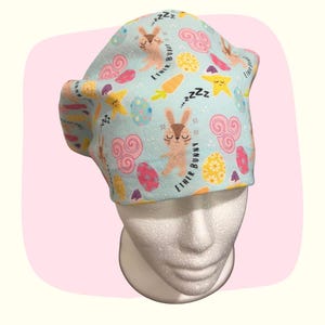 May include: A light blue patterned cap featuring a repeating design of sleeping bunnies, Easter eggs, carrots, and stars. The cap has the text "Ether Bunny" and "zzz" throughout the design. The cap is on a white mannequin head.