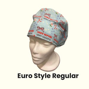 以下が含まれることがあります： 水色のEuro Style Regular手術用スクラブキャップ。赤いリボン、花、そして「VERY DEMURE」と「VERY MINDFUL」という言葉が赤色でパターン化されています。白いマネキンの頭にキャップが展示されています。
