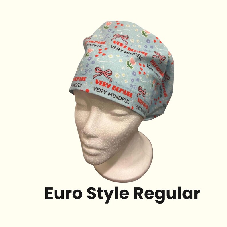以下が含まれることがあります： 水色のEuro Style Regular手術用スクラブキャップ。赤いリボン、花、そして「VERY DEMURE」と「VERY MINDFUL」という言葉が赤色でパターン化されています。白いマネキンの頭にキャップが展示されています。