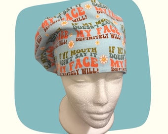 May include: A light blue scrub cap with the text "If my mouth doesn't say it, my face definitely will!" in orange and brown. The cap features a daisy pattern and is displayed on a white mannequin head.