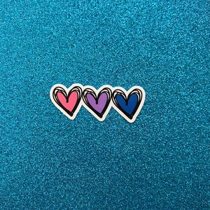 Puede incluir: Tres corazones dibujados a mano en rosa, morado y azul sobre un fondo de purpurina azul.