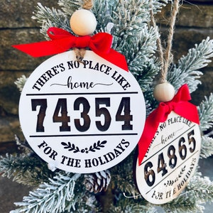 May include: Two white round wooden ornaments with red ribbon bows and a brown twine hanger. The ornaments have black text that reads "There's no place like home" and a house address number. The text below the address reads "For the Holidays".