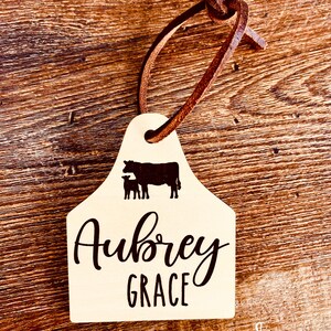 May include: A white wooden cow tag with a brown leather strap. The tag features a silhouette of a cow and calf with the text "Aubrey Grace" printed in black.