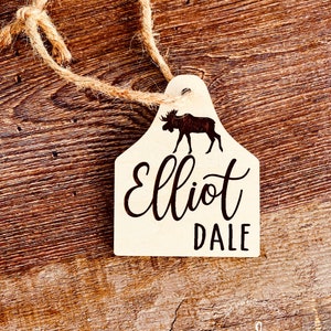 May include: Wooden ornament shaped like a moose head with the name "Elliot Dale" engraved on it. The ornament is attached to a twine string.