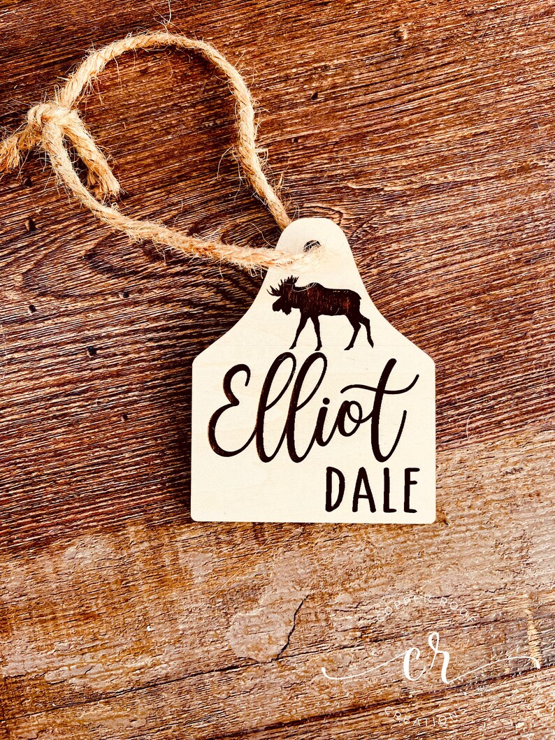 May include: Wooden ornament shaped like a moose head with the name "Elliot Dale" engraved on it. The ornament is attached to a twine string.