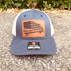 May include: A blue and white trucker hat with a brown leather patch featuring an American flag and the text "United we stand, disarmed we fall". The patch also includes a silhouette of a rifle. The hat is a Richardson 115 Low Pro Trucker.