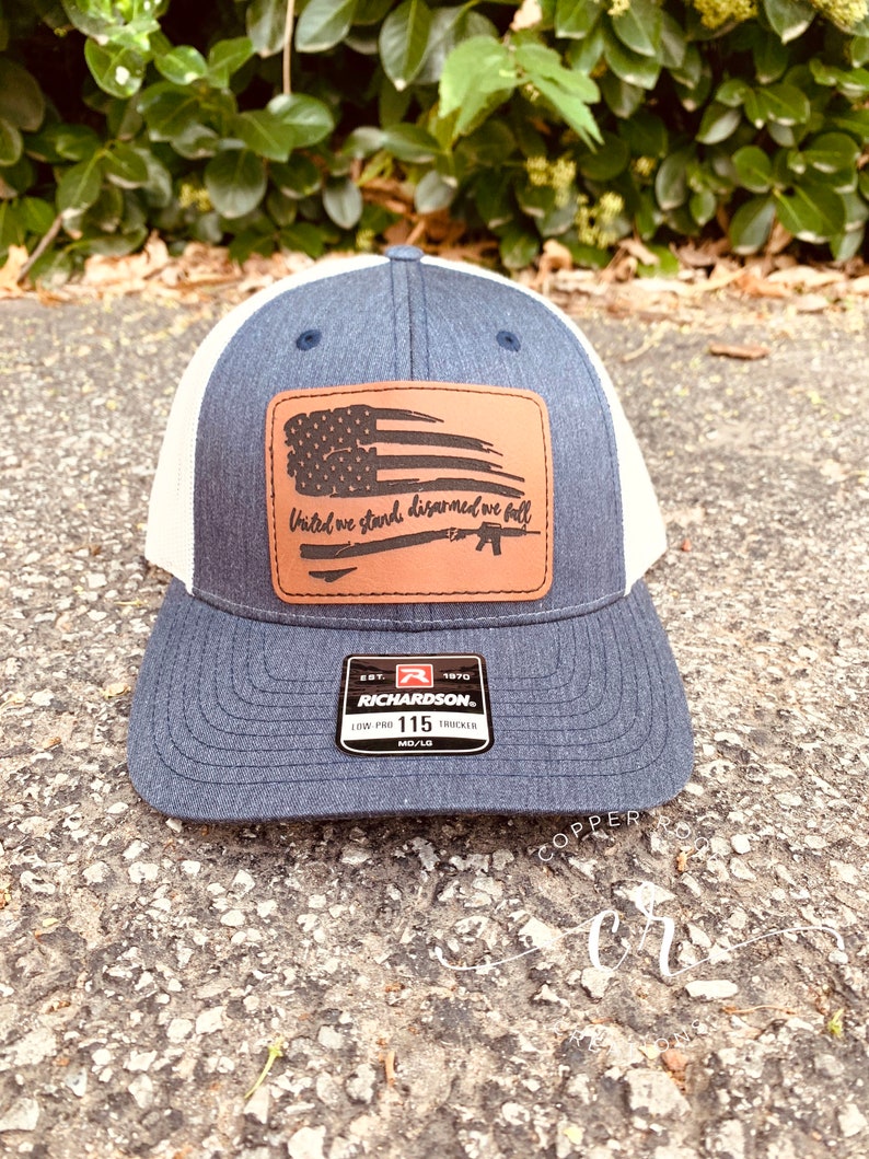 May include: A blue and white trucker hat with a brown leather patch featuring an American flag and the text "United we stand, disarmed we fall". The patch also includes a silhouette of a rifle. The hat is a Richardson 115 Low Pro Trucker.