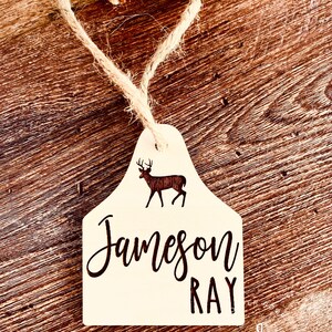 May include: A white wooden deer tag with a silhouette of a deer and the text "Jameson Ray" in black script.