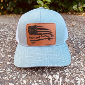 May include: A gray and white trucker hat with a brown leather patch featuring a distressed American flag and the text "United we stand, disarmed we fall".