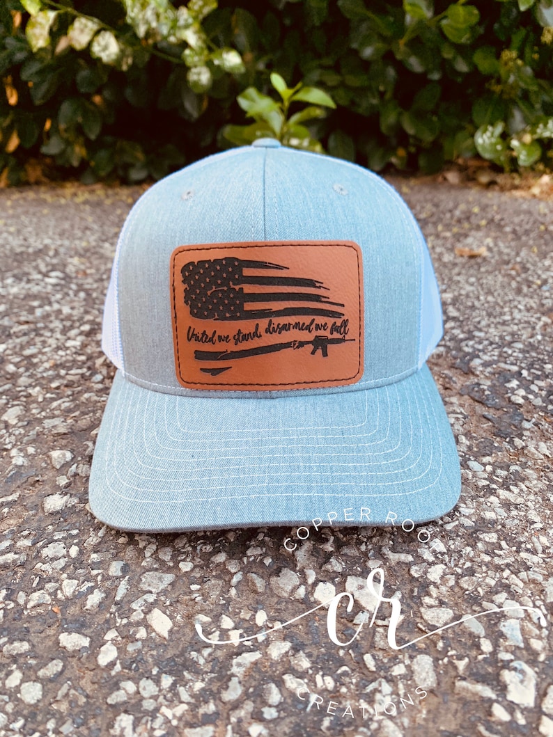May include: A gray and white trucker hat with a brown leather patch featuring a distressed American flag and the text "United we stand, disarmed we fall".