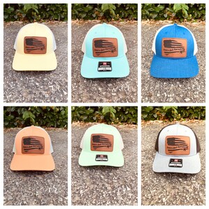 May include: Six different colored trucker hats with a white mesh back. Each hat has a brown leather patch with an American flag and the text "Hold on, God doesn't make mistakes".