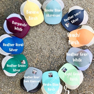 May include: A variety of trucker hats in different colors and patterns. The hats are arranged in a circle on a gray surface. The hats have different color combinations, including burgundy and white, heather royal and silver, smoke blue and aluminum, heather navy and silver, peach and birch, heather gray and white, heather green and silver, heather gray and dark charcoal, light green and birch, and aruba blue and birch.