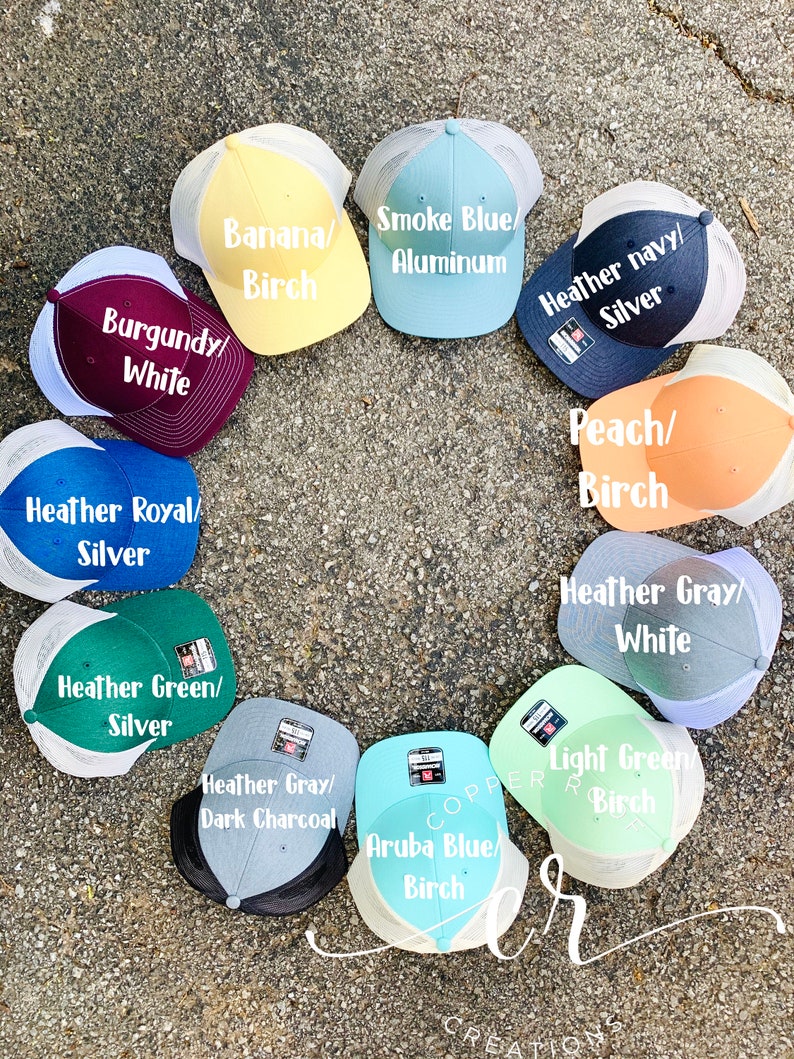 May include: A variety of trucker hats in different colors and patterns. The hats are arranged in a circle on a gray surface. The hats have different color combinations, including burgundy and white, heather royal and silver, smoke blue and aluminum, heather navy and silver, peach and birch, heather gray and white, heather green and silver, heather gray and dark charcoal, light green and birch, and aruba blue and birch.