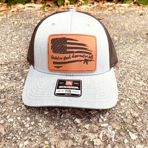 May include: A gray and black trucker hat with a brown leather patch that says "United we stand, disarmed we fall" with a silhouette of an American flag and a rifle. The hat has a Richardson 115 label.