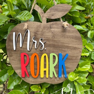May include: Wooden apple-shaped sign with the text "Mrs. Roark" in rainbow colors. The sign is hanging from a twine string and has a brown wood grain finish.