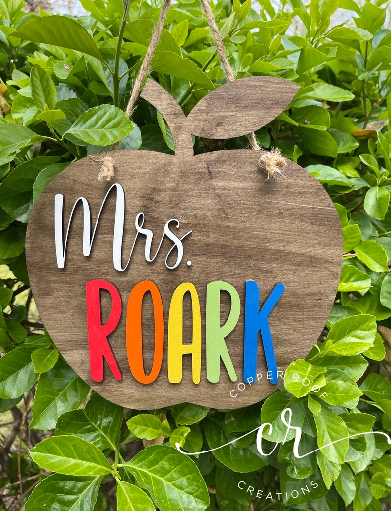May include: Wooden apple-shaped sign with the text "Mrs. Roark" in rainbow colors. The sign is hanging from a twine string and has a brown wood grain finish.