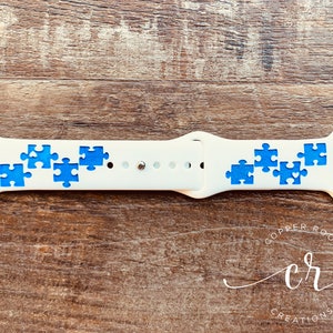 May include: White Apple Watch band with blue puzzle pieces printed on it. The band is made of silicone and has a buckle closure.