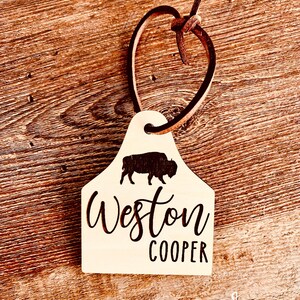 May include: A wooden tag with a brown leather cord. The tag is shaped like a cow tag and has a silhouette of a buffalo. The tag is engraved with the text "Weston Cooper".