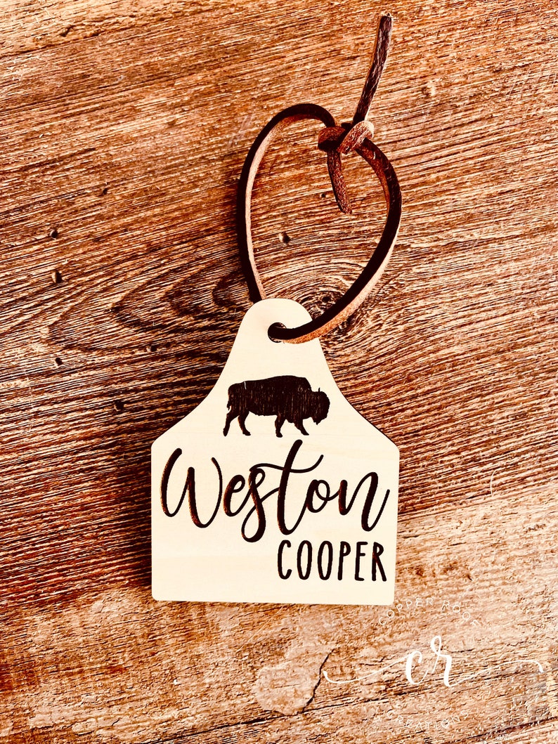 May include: A wooden tag with a brown leather cord. The tag is shaped like a cow tag and has a silhouette of a buffalo. The tag is engraved with the text "Weston Cooper".