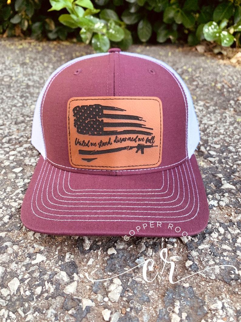 May include: A maroon and white trucker hat with a brown leather patch featuring a distressed American flag and the text "United we stand, disarmed we fall".