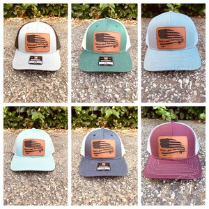 May include: Six trucker hats with a white mesh back and a front panel in different colors. The front panel features a brown leather patch with a distressed American flag design and the text "Hold on, God doesn't make mistakes". The hats are available in gray, green, light blue, navy blue, burgundy, and light blue.